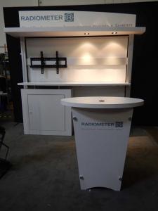 RENTAL: RE-1019 with White Laminated Sconce with Puck Lighting, RE-1201 Tapered Counter, White Laminated Backwall Counter & Shelves, Large Monitor Mount, 42" Monitor, RE-171 Literature Stand, Tension Fabric Panel for Backwall, Sintra Header Graphic, and V