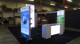 Modified ECO-4094 Sustainable Island with LED Backlit Fabric Graphics, Locking Closet, Double-sided Lightboxes with Shelves or Roller Bars, Curved  Headers, Monitor Mount, and ECO-42C Backlit Counter with Storage